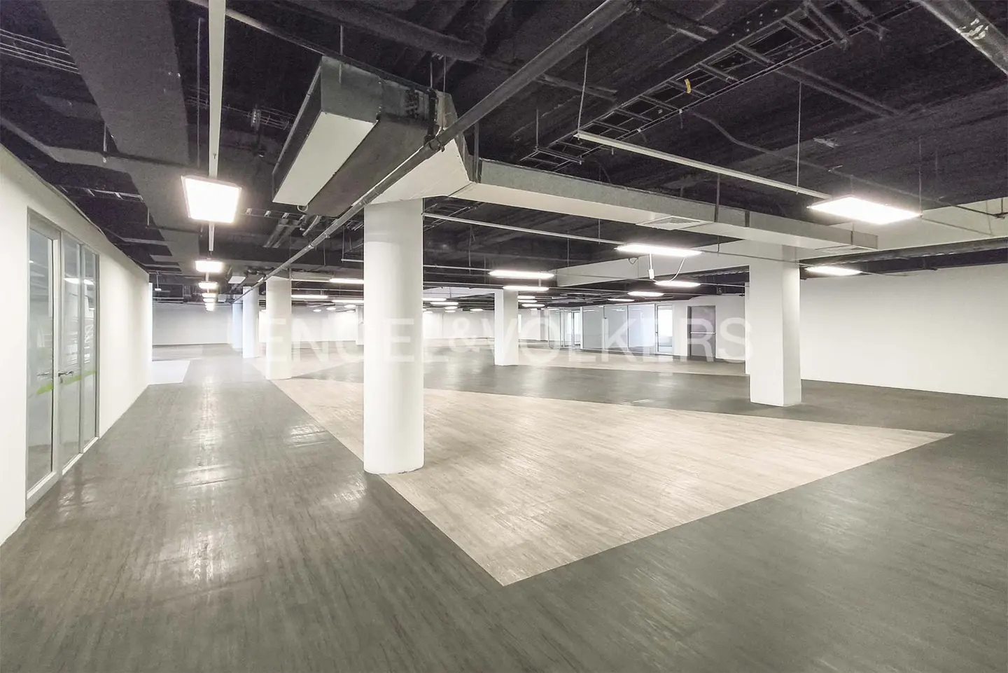 Bright, empty commercial space with gray and light wood floors, white walls, and white support columns. Black ceiling with exposed pipes and lighting.