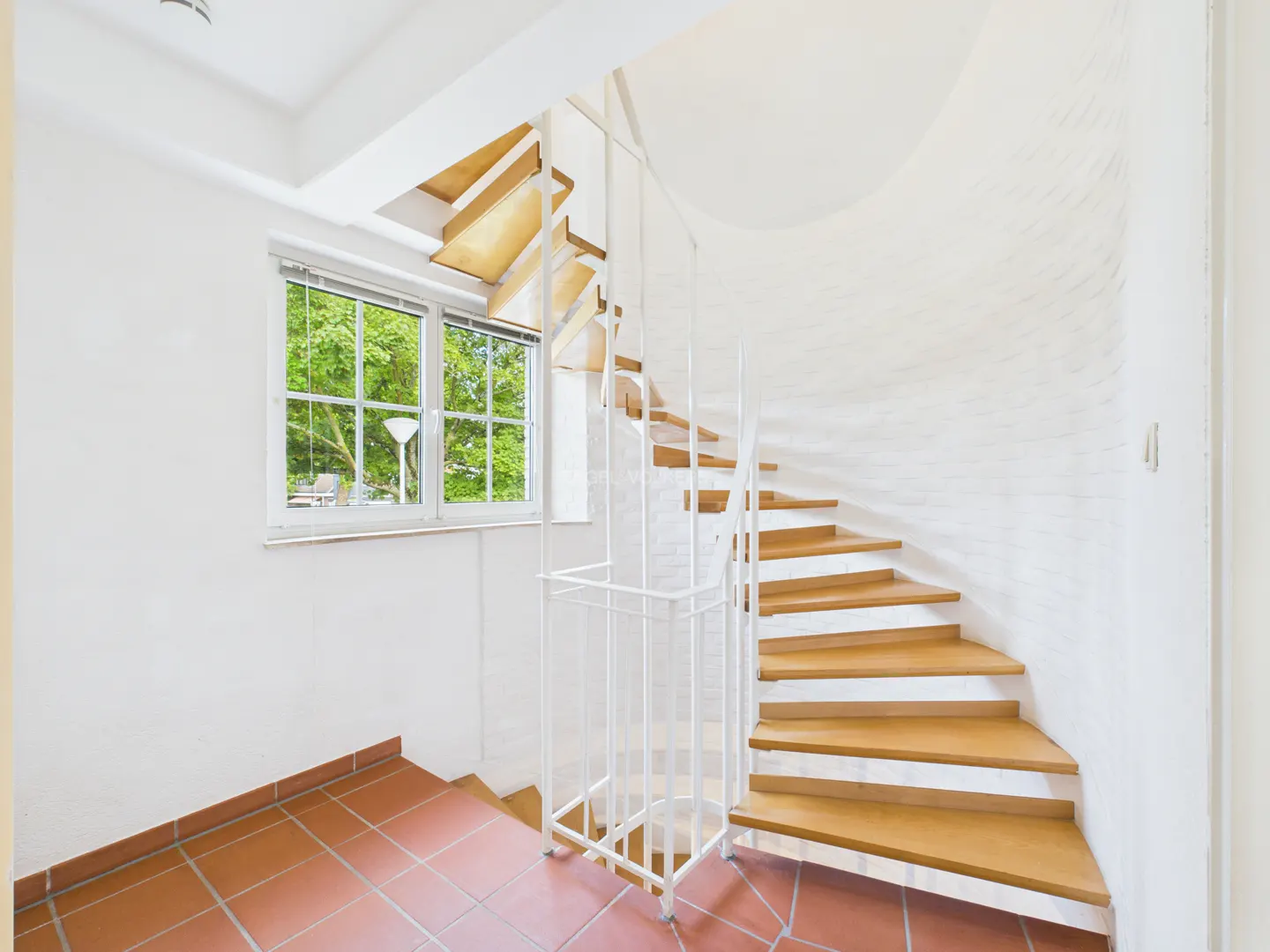 A bright room with a wooden spiral staircase and a white metal railing. A window shows green trees outside. The floor is tiled in red.