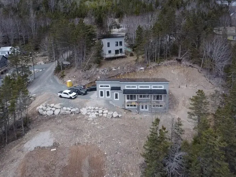Aerial view of a gray, modern house on a hill surrounded by trees. Cars are parked nearby, and a second house is visible in the background.