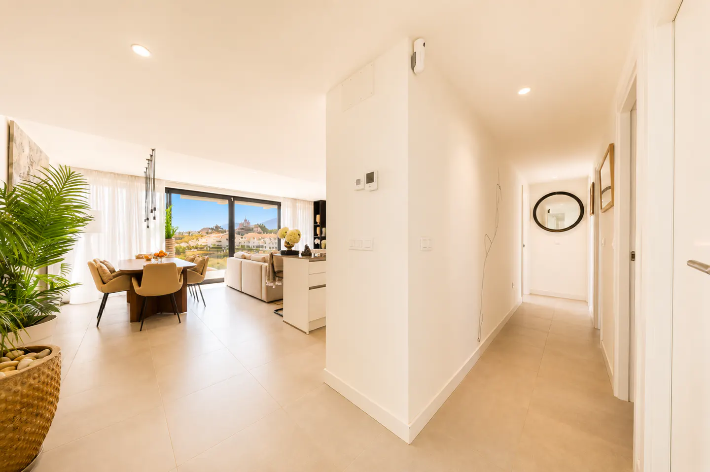 Bright, modern apartment interior with dining table, tan chairs, and a large window view. A hallway leads to other rooms. A potted plant is in the foreground.