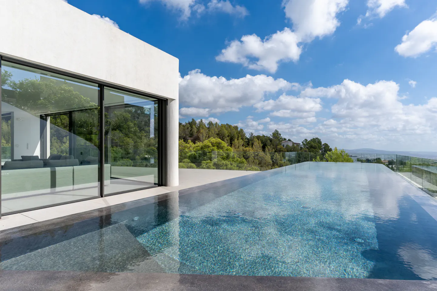 Modern white house with an infinity pool overlooking trees and a blue sky with white clouds. Large glass doors reflect the interior.