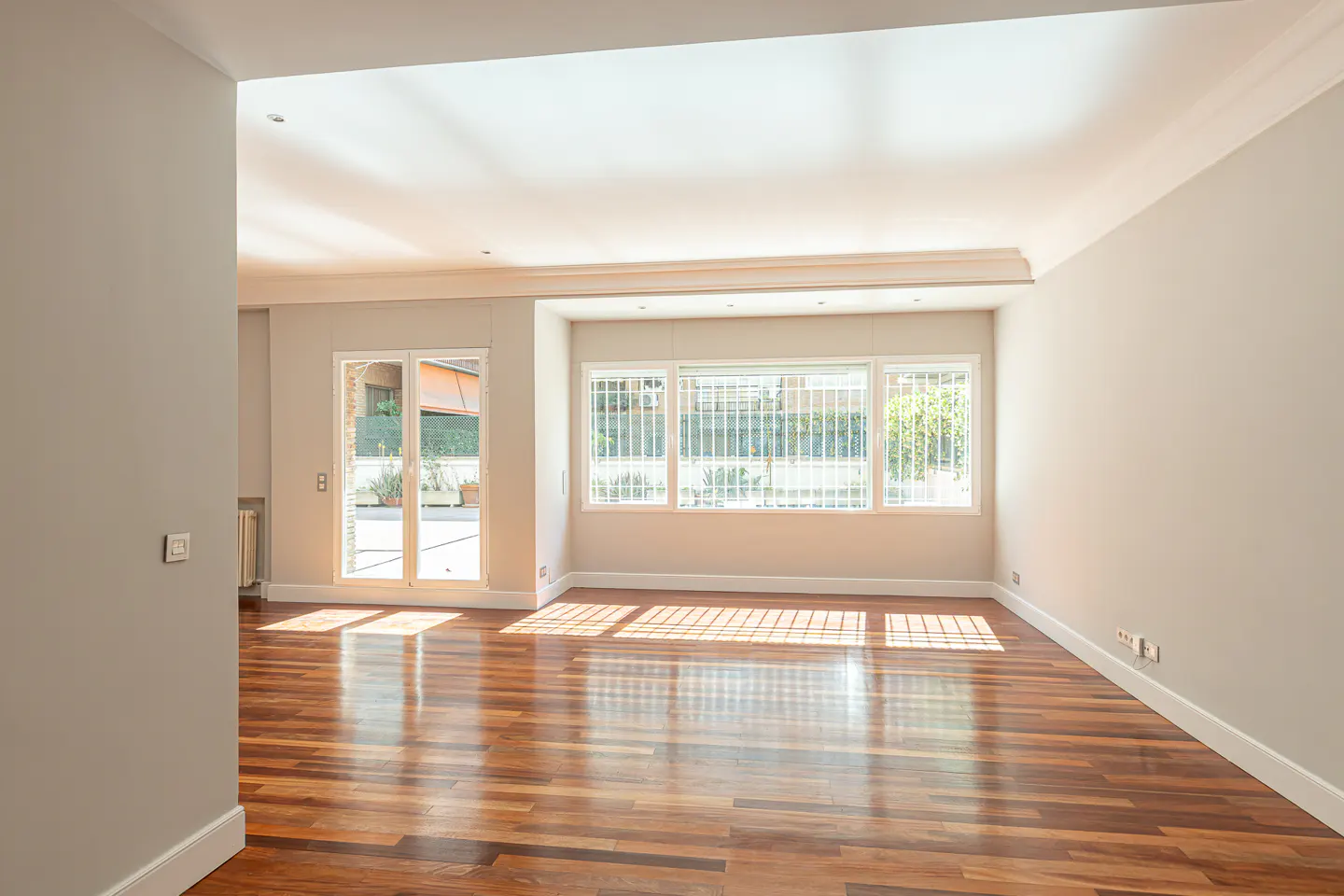 Bright, empty room with hardwood floors, white walls, and large windows with white bars. Sunlight streams in, creating shadows on the floor.
