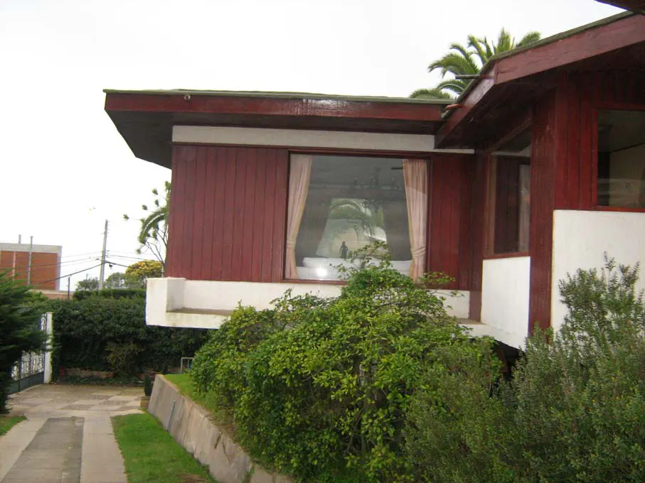 Exterior view of a red wood house with a large window and green bushes in front.