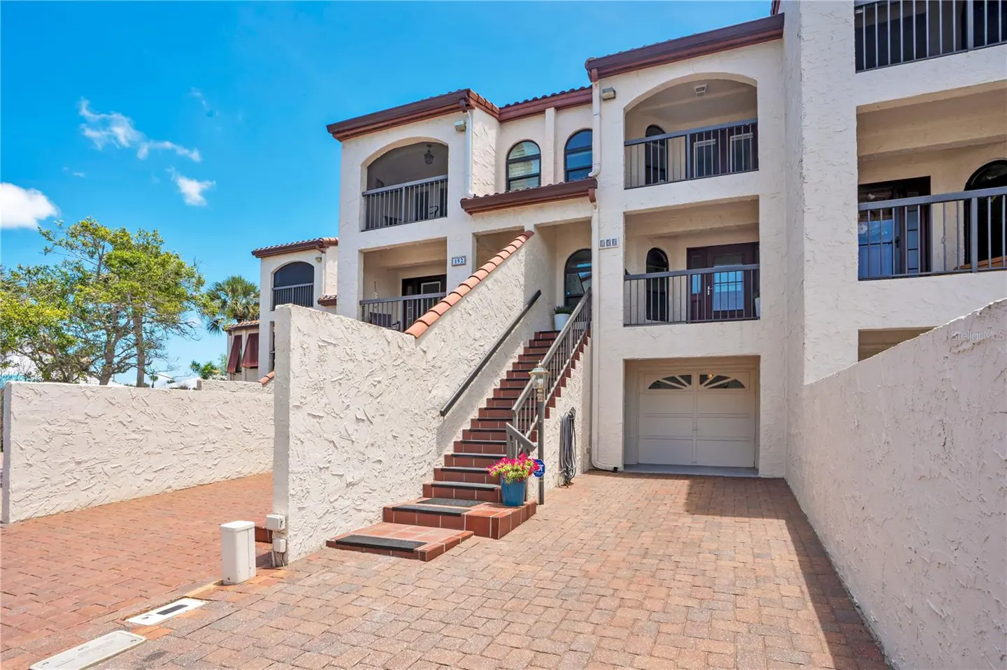 Exterior of a multi-story cream stucco condo building with arched balconies, a brick driveway, and stairs to the entrance.
