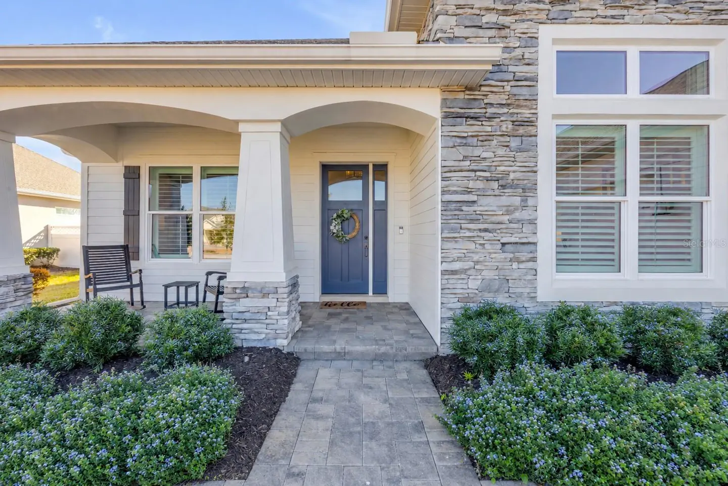 Home exterior featuring a blue front door with a wreath, stone accents, and a paved walkway lined with green bushes.