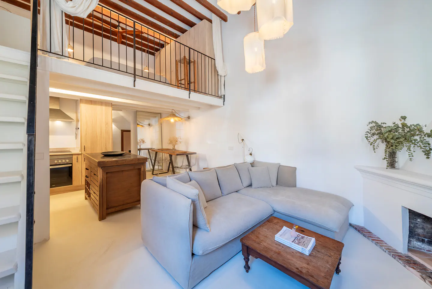 Bright, open-plan living space with a loft. A light gray sectional sofa faces a wooden coffee table. Kitchen area with island and dining table visible. White walls and wooden beams.