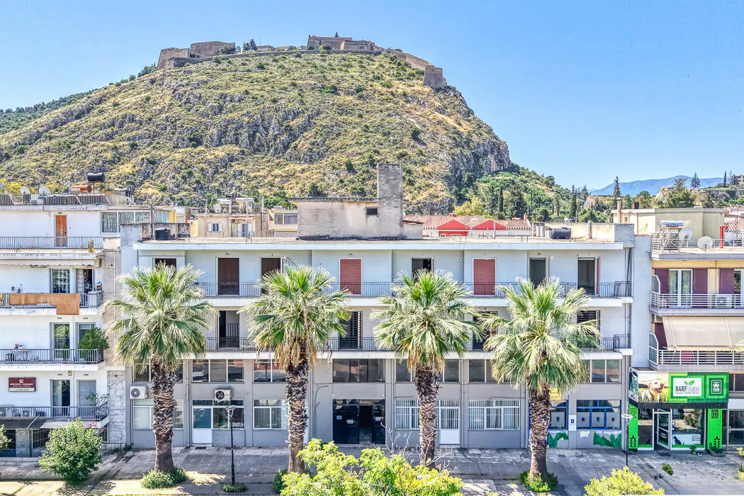 Cityscape view of a building with palm trees in front and a mountain with a fortress in the background.