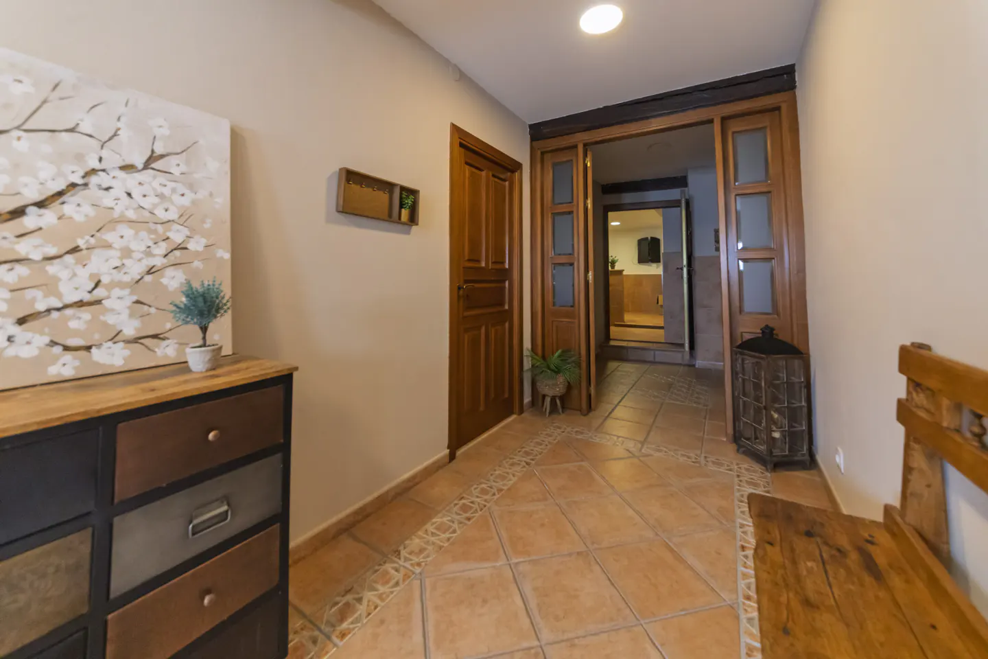 Hallway with terracotta tile floor, wooden doors, and a cabinet with a floral painting above it.