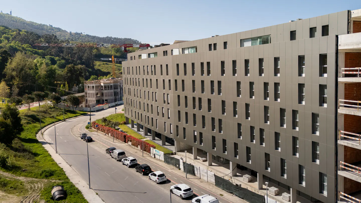 Modern gray apartment building under construction next to a road with parked cars and a green hillside in the background.