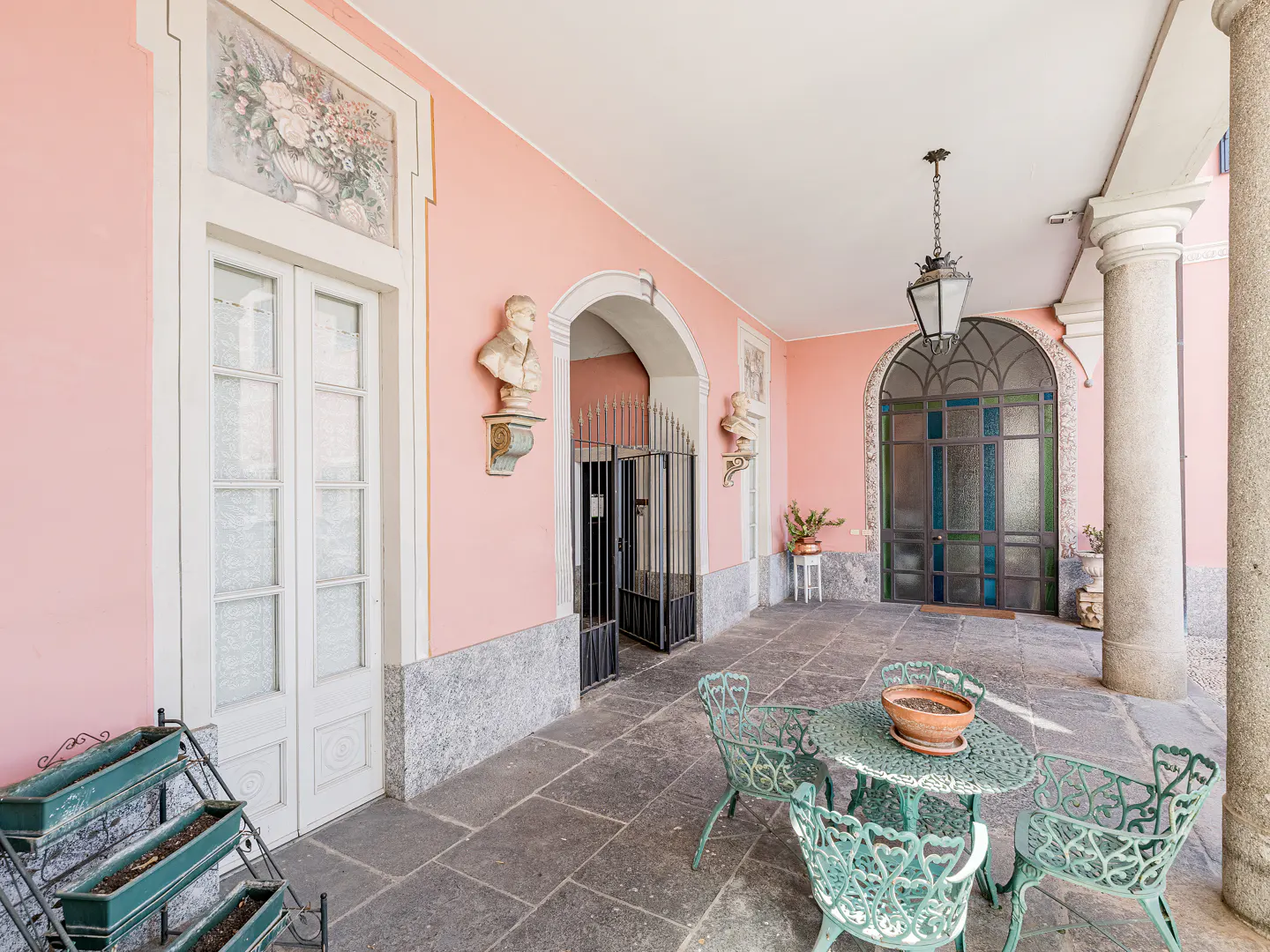 Covered patio with pink walls, stone floors, and white columns. A green metal table and chairs sit in the foreground. Busts adorn the walls.
