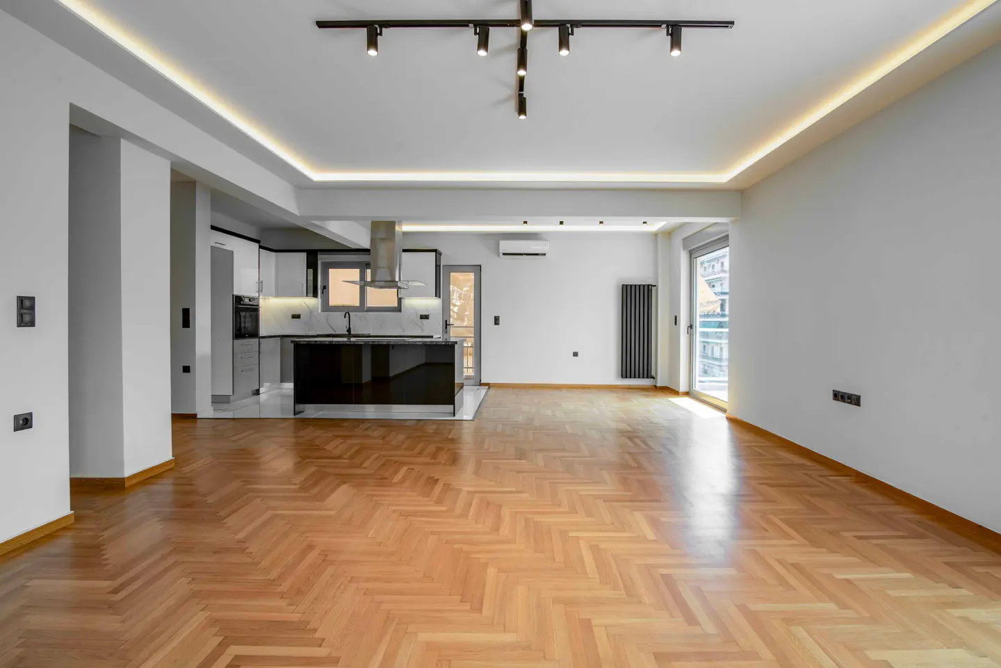 Bright, open-concept apartment with herringbone wood floors, white walls, and a modern kitchen with a black island. Track lighting on the ceiling.