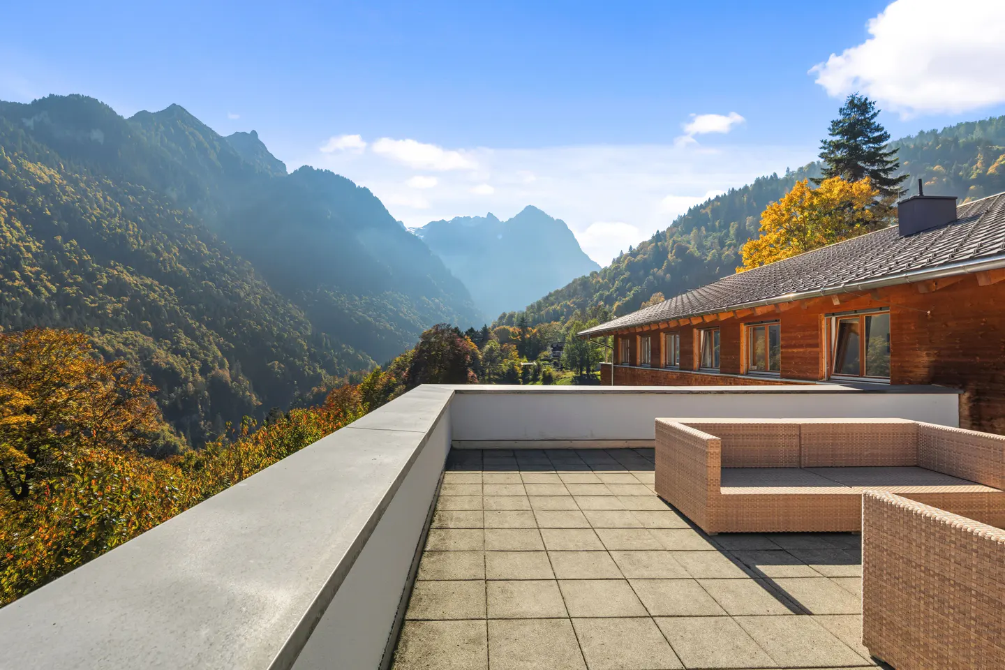 Scenic view from a patio with wicker furniture, overlooking mountains and trees under a blue sky.
