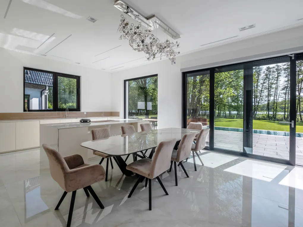 Bright, modern dining room with a marble table, tan chairs, and a unique chandelier. Sliding glass doors open to a green lawn and pool.
