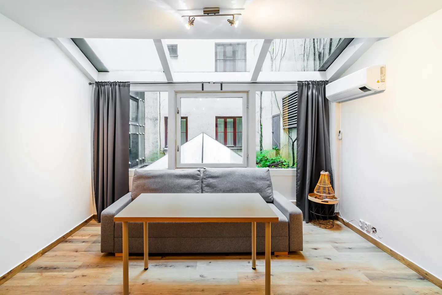 Modern room with a grey sofa, wooden table, and large skylight windows looking out onto a courtyard with grey curtains.
