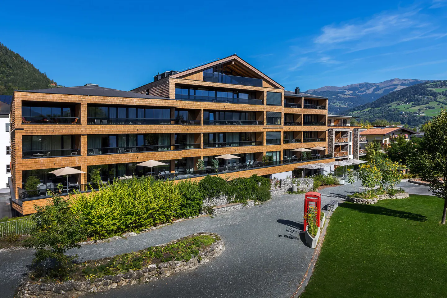 Exterior view of a modern, multi-story hotel with wood facade, balconies, and mountain backdrop on a sunny day.