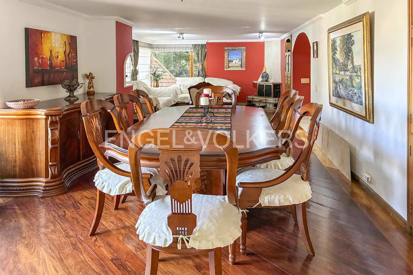 Formal dining room with a long wooden table, carved chairs with white cushions, red accent walls, and hardwood floors.
