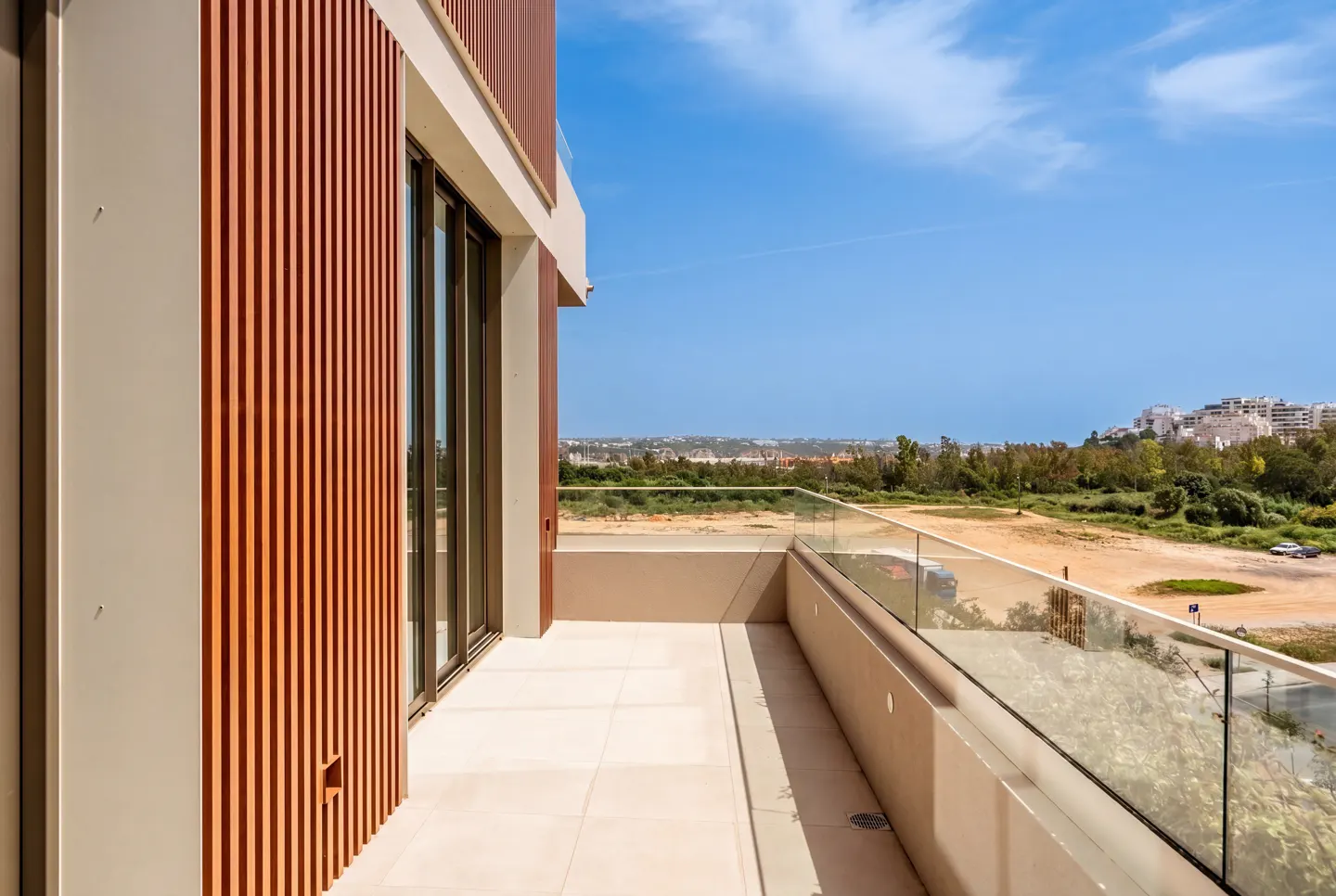 Balcony view with glass railing, beige tile floor, and wood-slatted wall. Distant buildings and blue sky visible.