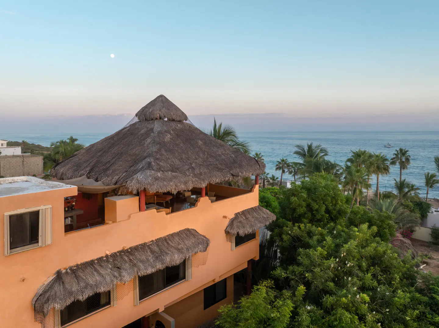 A peach-colored building with a thatched roof overlooks the ocean at dusk. Palm trees and lush greenery surround the building.