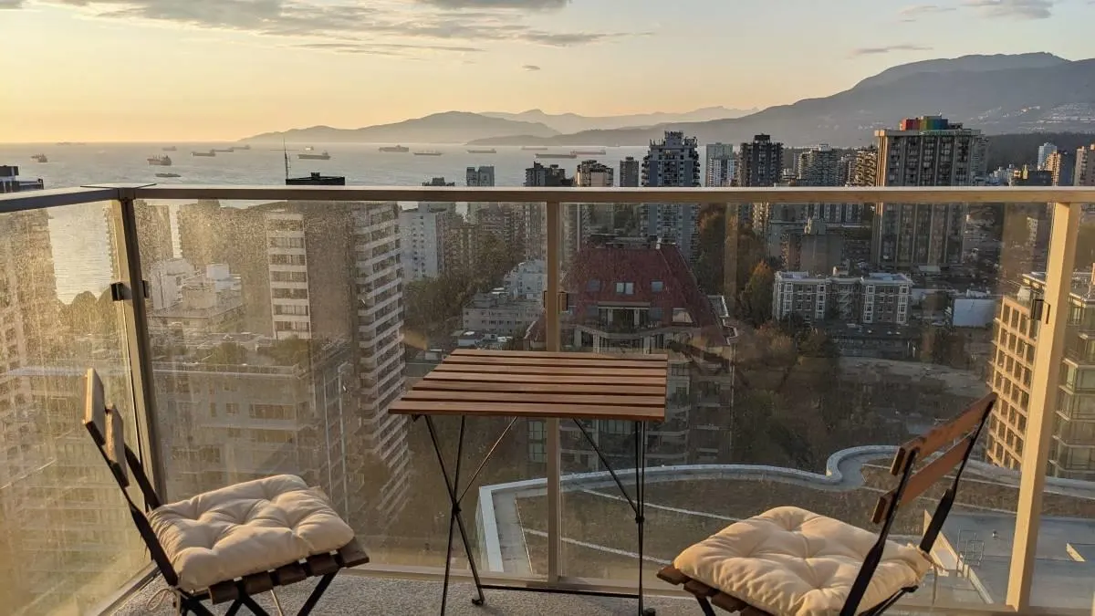 Balcony view at sunset. Table and chairs overlook city buildings, ocean with ships, and distant mountains.