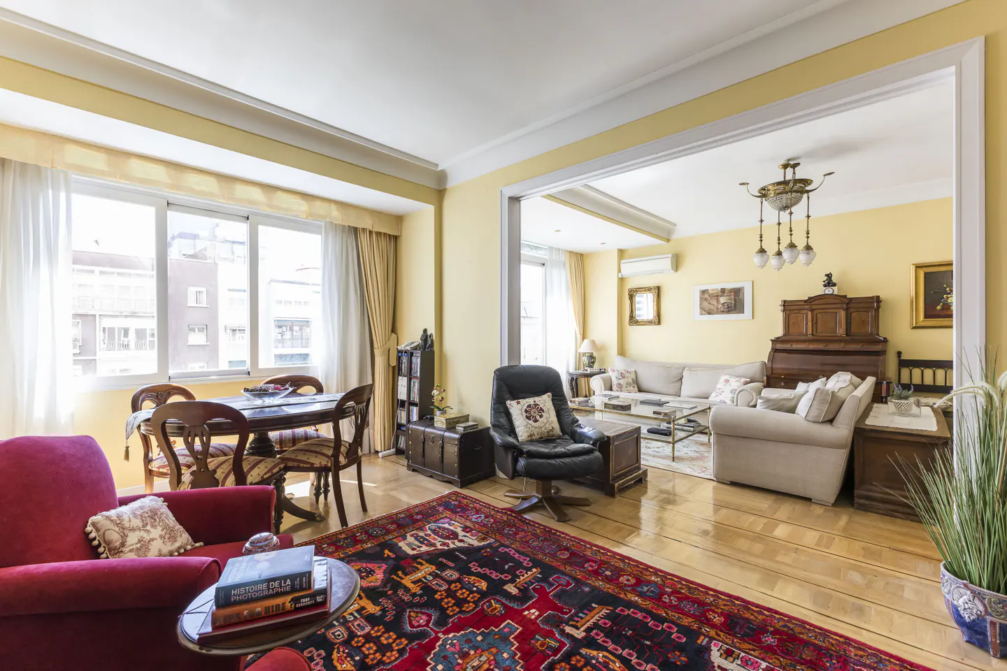 A bright living room with yellow walls, hardwood floors, and a red patterned rug. A red armchair and dining table are visible in the foreground.