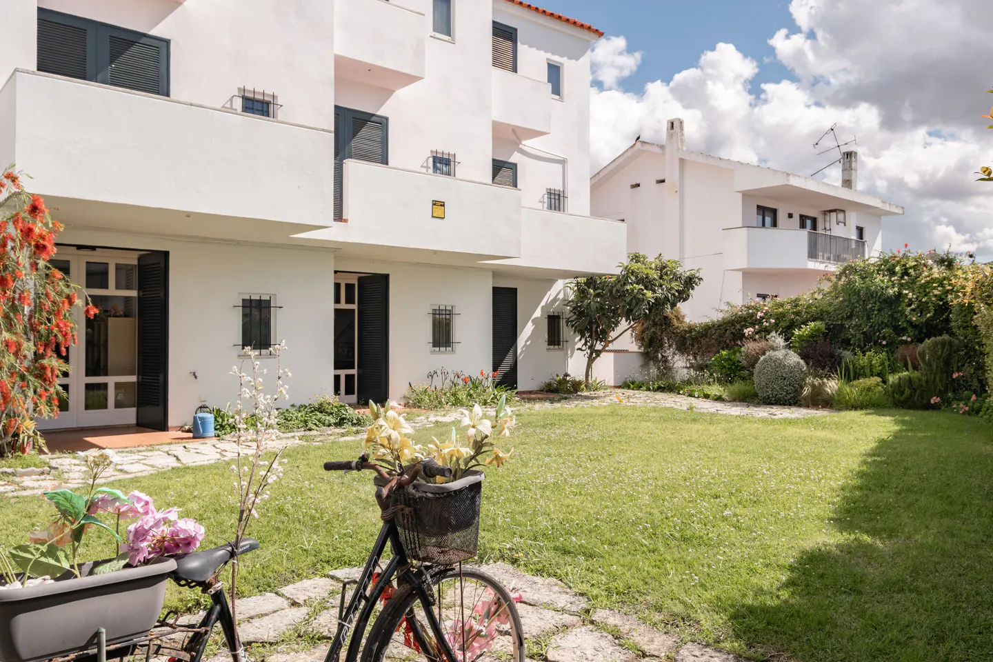 A white building with black shutters overlooks a green lawn. A bicycle with flowers in the basket sits on a stone path.