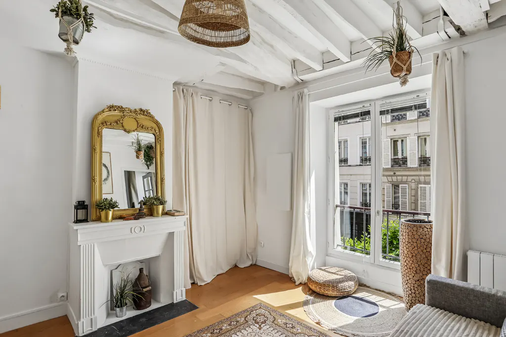 Bright living room with white walls, exposed beams, and a large window showing a city view. A gold mirror sits above a white fireplace.