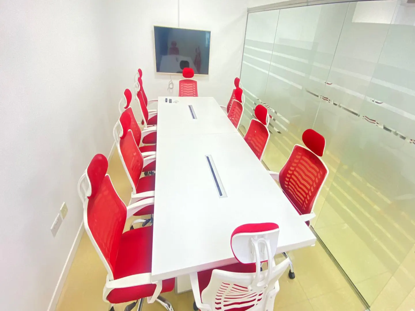 Bright conference room with a long white table, red mesh chairs, and a wall-mounted TV. A glass wall adds natural light.