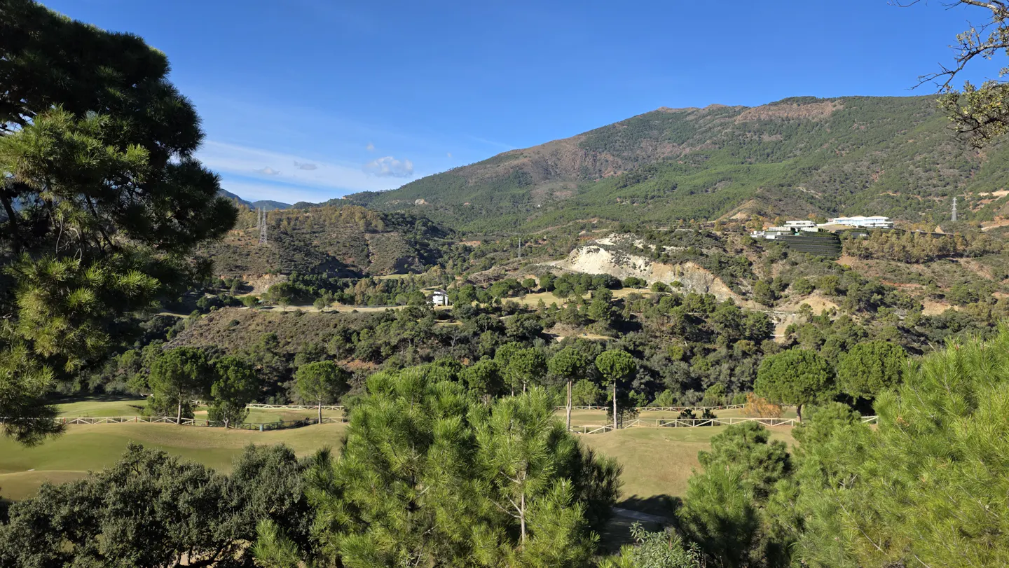 Scenic view of a lush green golf course nestled in a valley surrounded by pine-covered mountains under a clear blue sky.