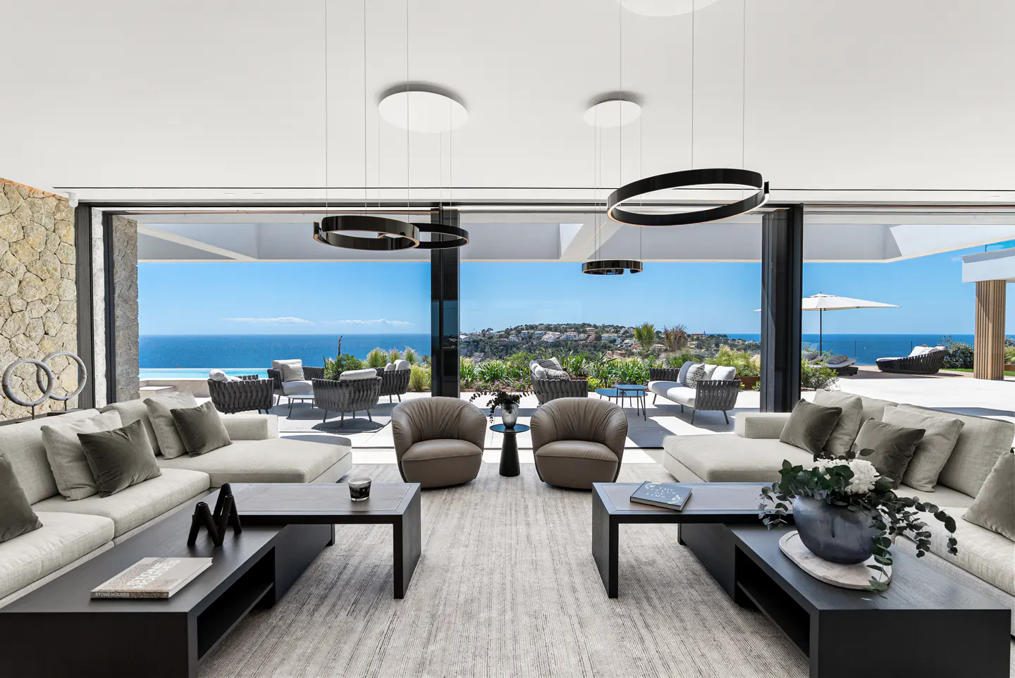 Bright living room with sofas, chairs, and black coffee tables. Large sliding doors open to a patio with ocean views.