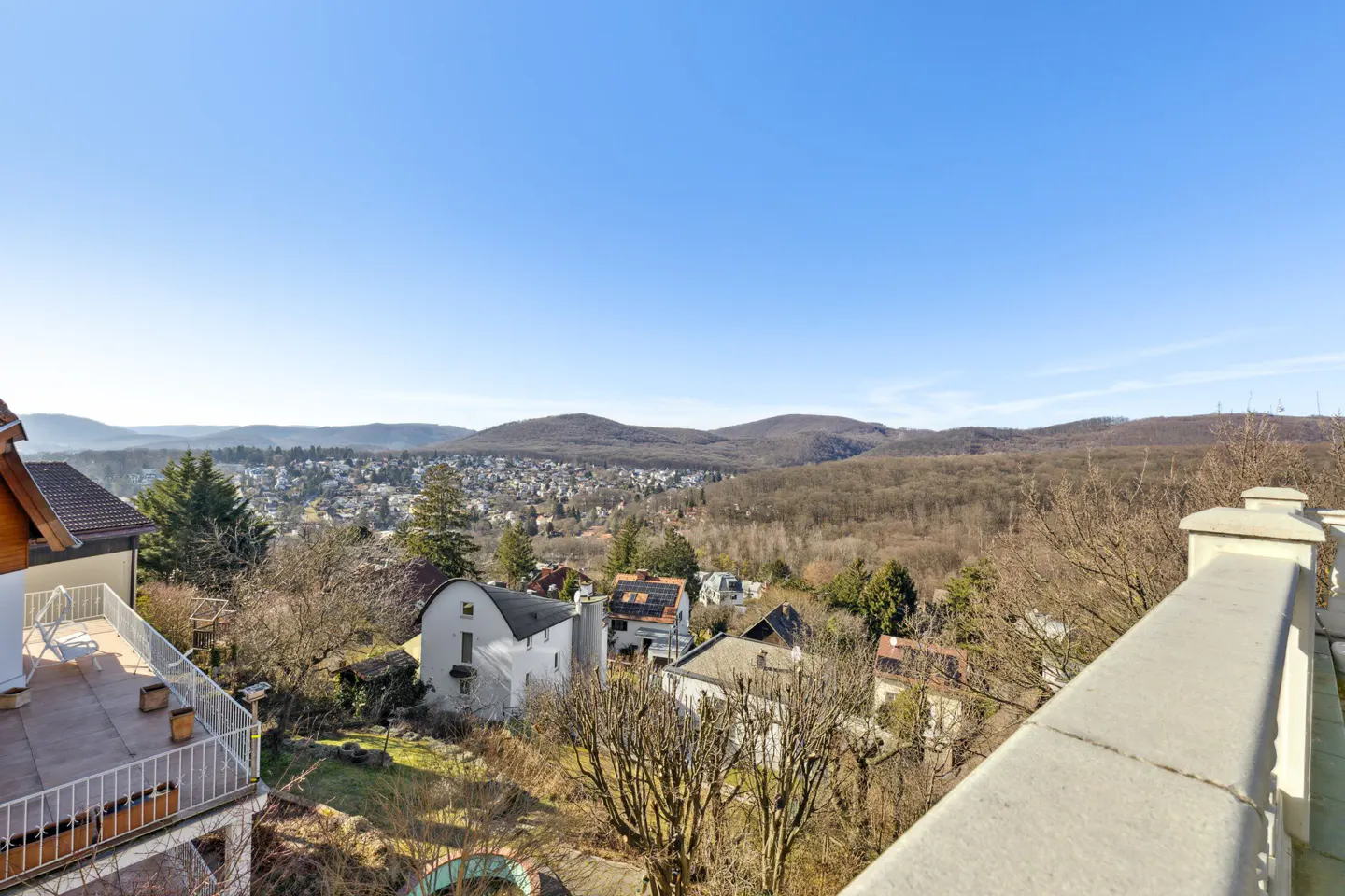 View from a balcony overlooking houses, trees, and hills under a clear blue sky.