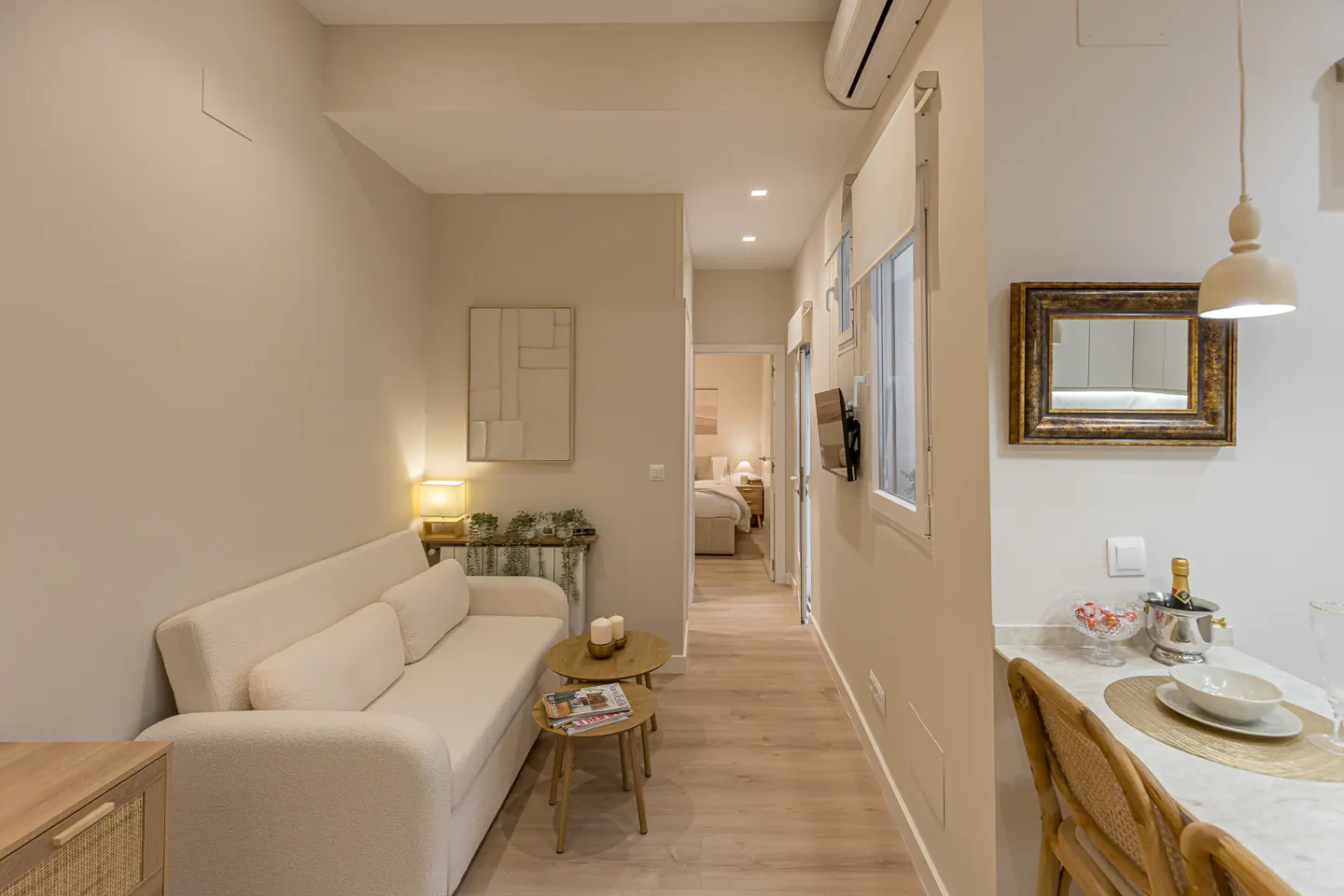 A bright, narrow apartment interior with a white sofa, wood floors, and a glimpse of a bedroom in the background.