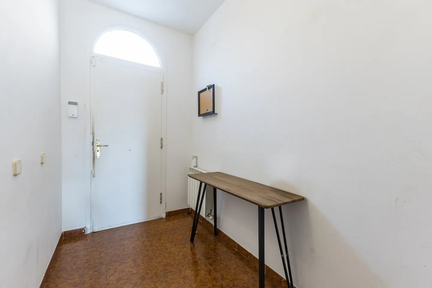 Entryway with white walls, brown tile floor, and a wooden table with black legs. A white door with a rounded top is on the left.