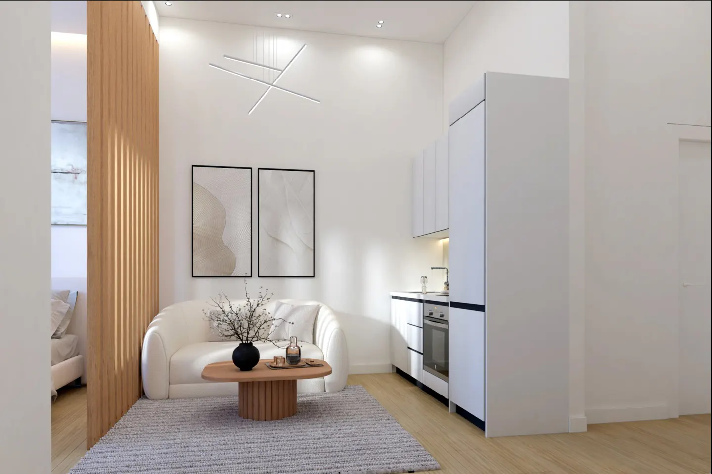 Bright, modern apartment interior with white walls, wood floors, and a gray rug. A white sofa faces a round wood table with a black vase. A small kitchen is visible.