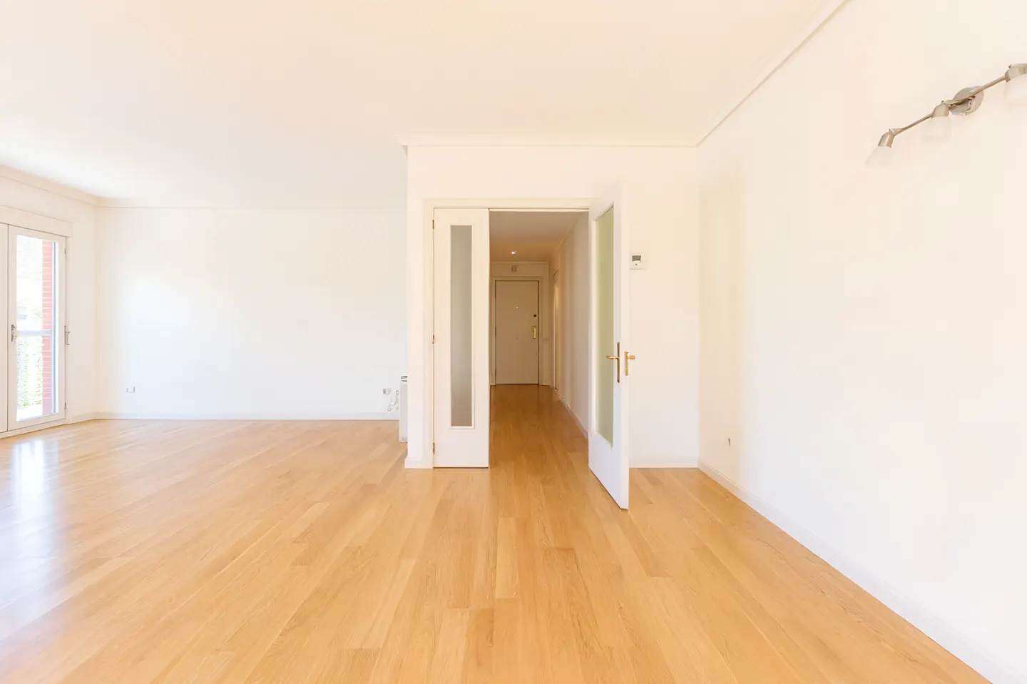 Bright, empty room with light wood floors and white walls. French doors lead to a balcony. Glass paneled doors open to a hallway.