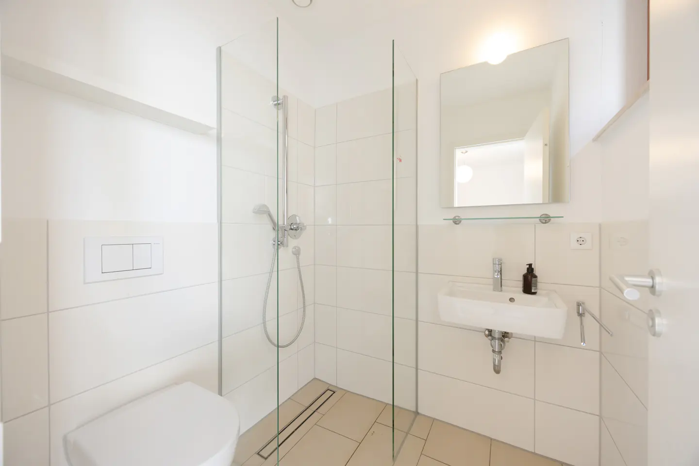 Bright bathroom with white tiles, a glass-enclosed shower, a toilet, and a sink with a mirror above it.