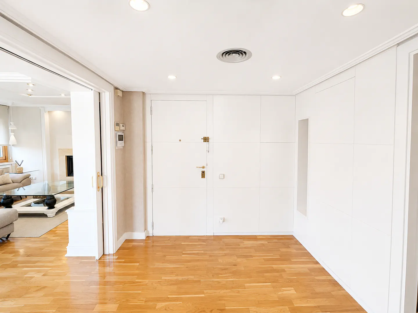 Bright, empty room with light wood floors, white walls, and recessed lighting. A doorway leads to a living room with a fireplace.