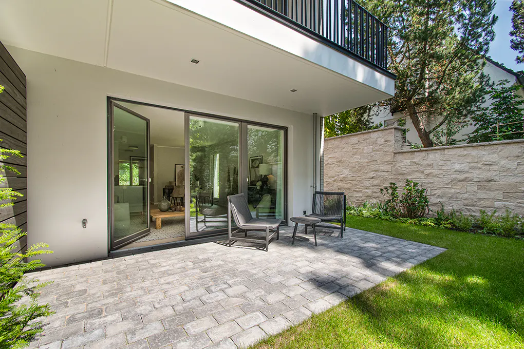 Modern home exterior with gray patio, chairs, and open sliding glass doors. A stone wall and green lawn are in the background.