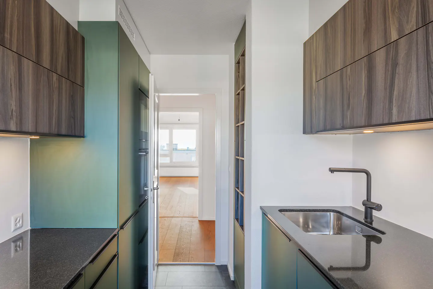 A modern kitchen with green cabinets, dark wood upper cabinets, and a black countertop with a stainless steel sink. A doorway leads to a bright room.