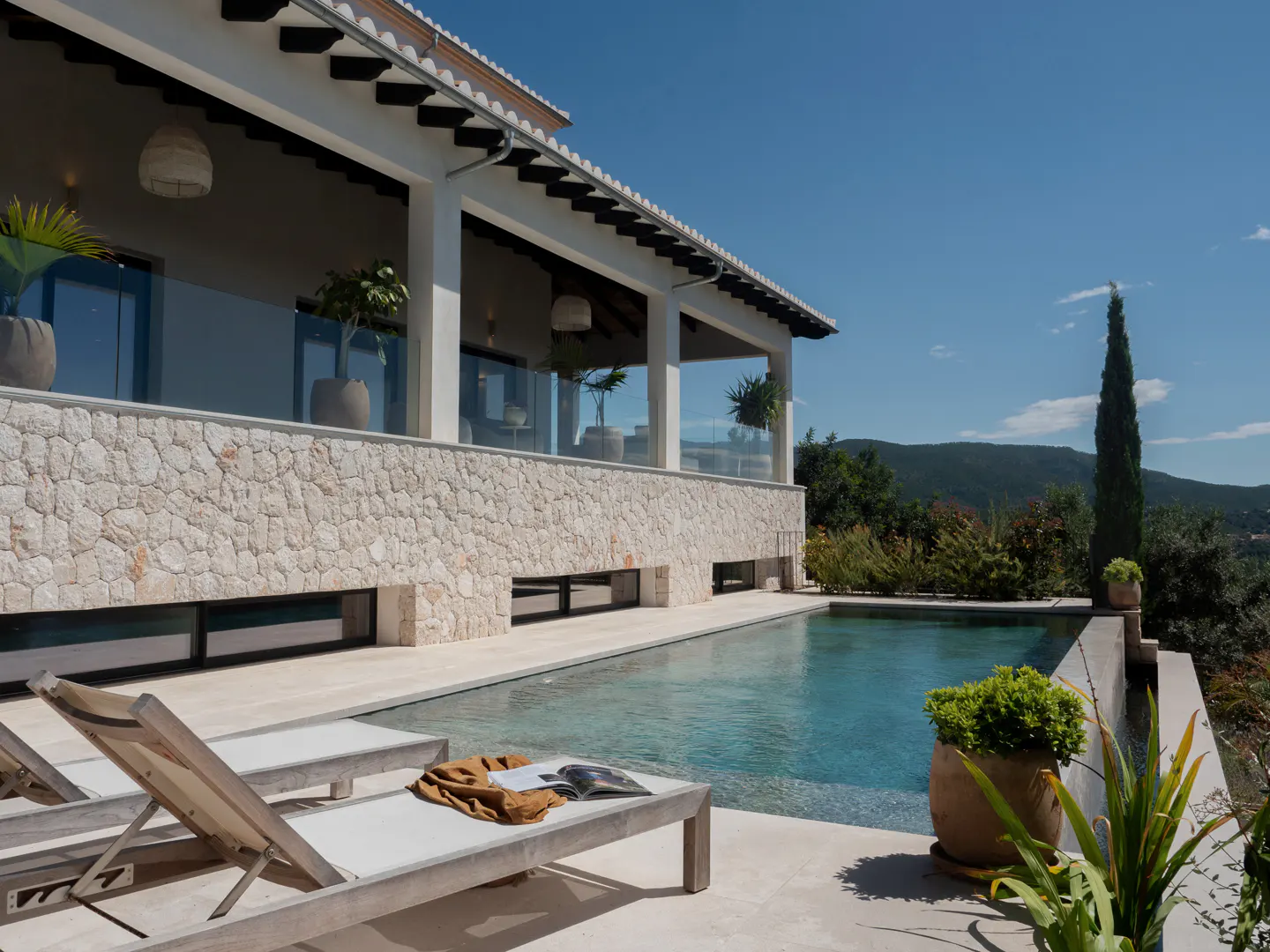 Exterior view of a luxury villa with a pool, stone facade, lounge chairs, and a scenic mountain backdrop under a clear blue sky.
