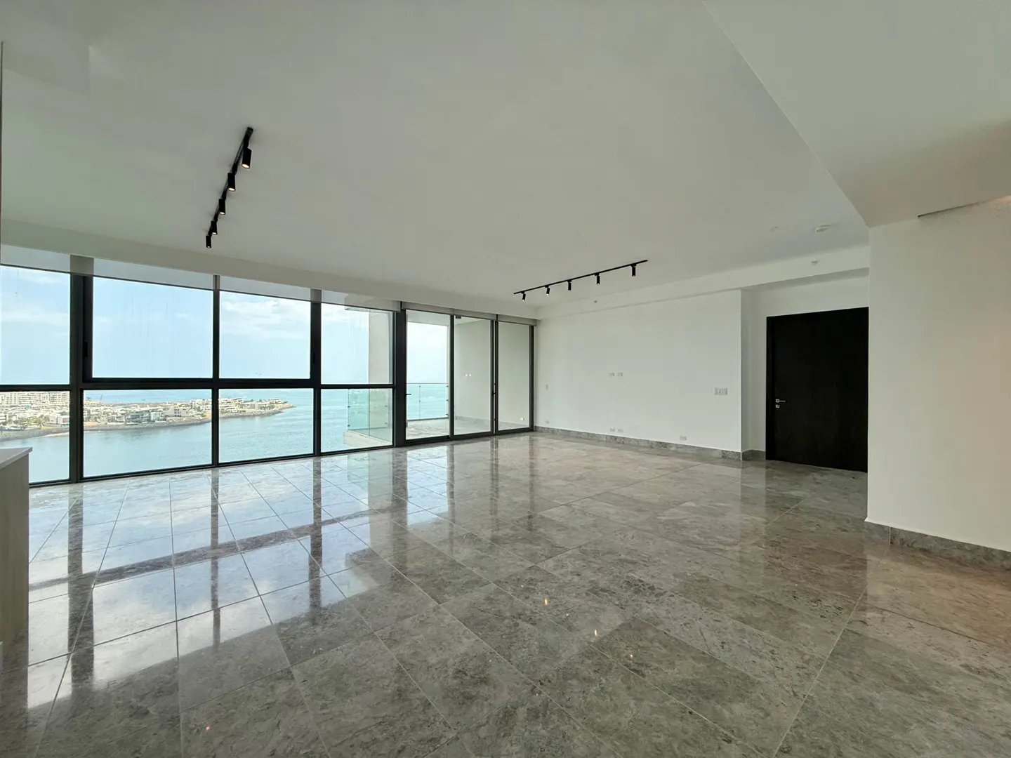 Empty, bright room with gray tile floor, white walls, and floor-to-ceiling windows overlooking the ocean. Black track lighting on the ceiling.
