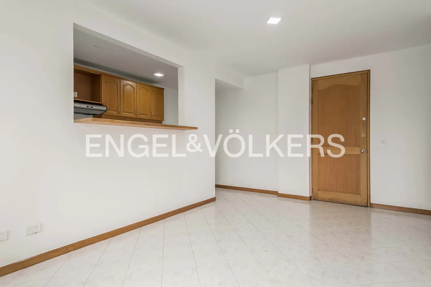 Bright room with white walls, light tile floors, a wooden door, and a kitchen pass-through window with wooden cabinets.