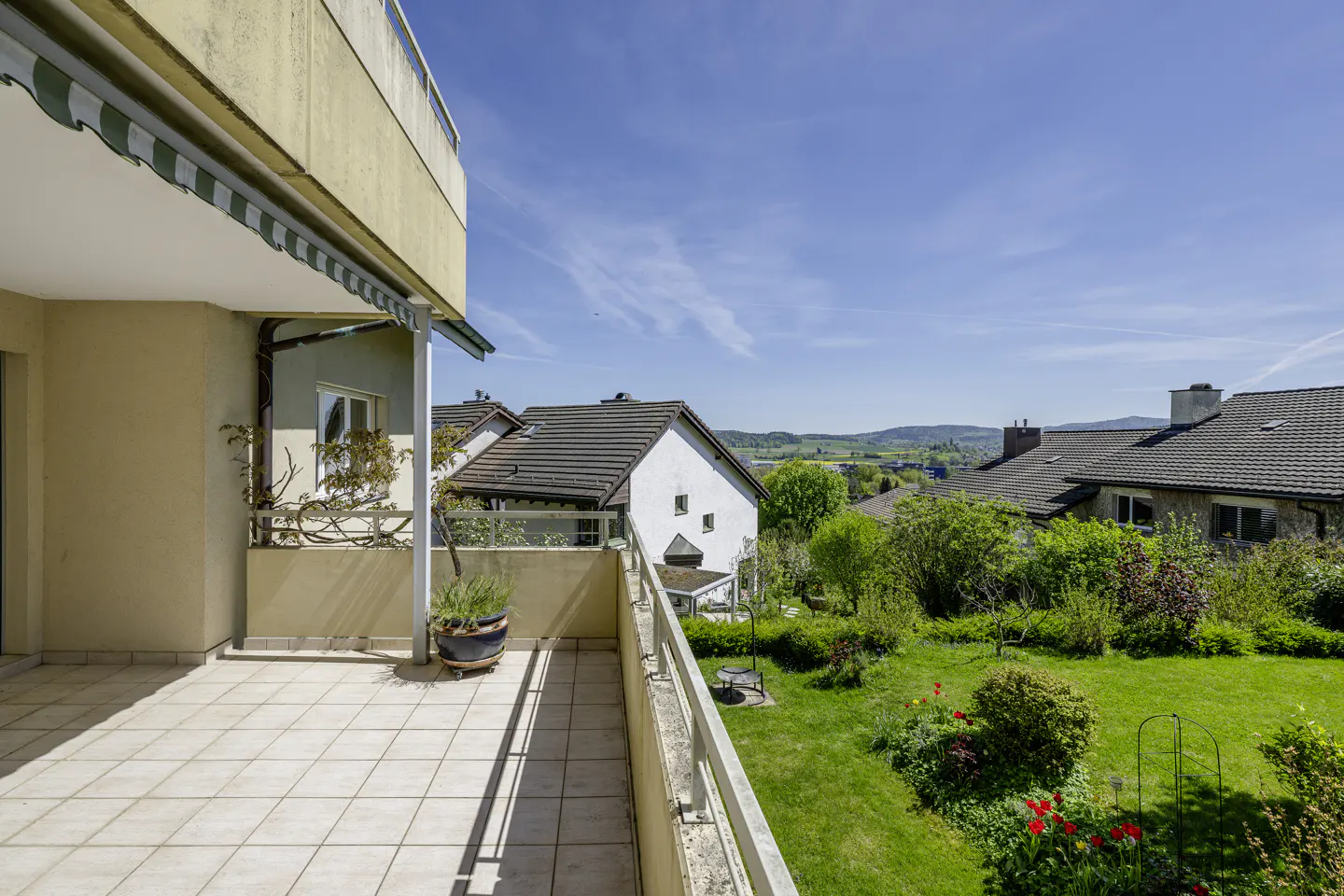 Spacious tiled balcony overlooking a lush green garden, neighboring houses, and distant hills under a clear blue sky.