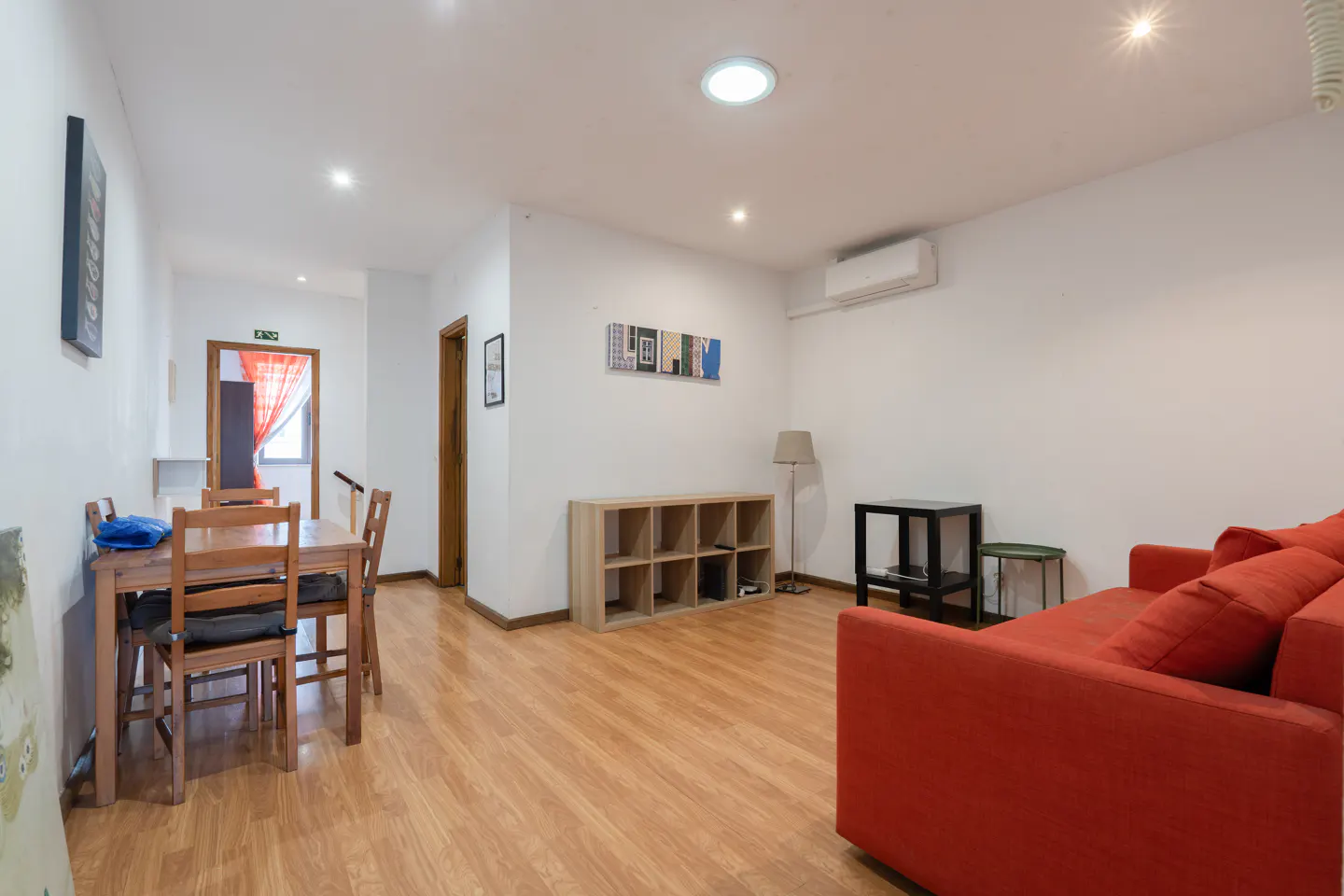 Bright living room with wood floors, white walls, and recessed lighting. A red sofa, dining table, and shelving unit furnish the space.