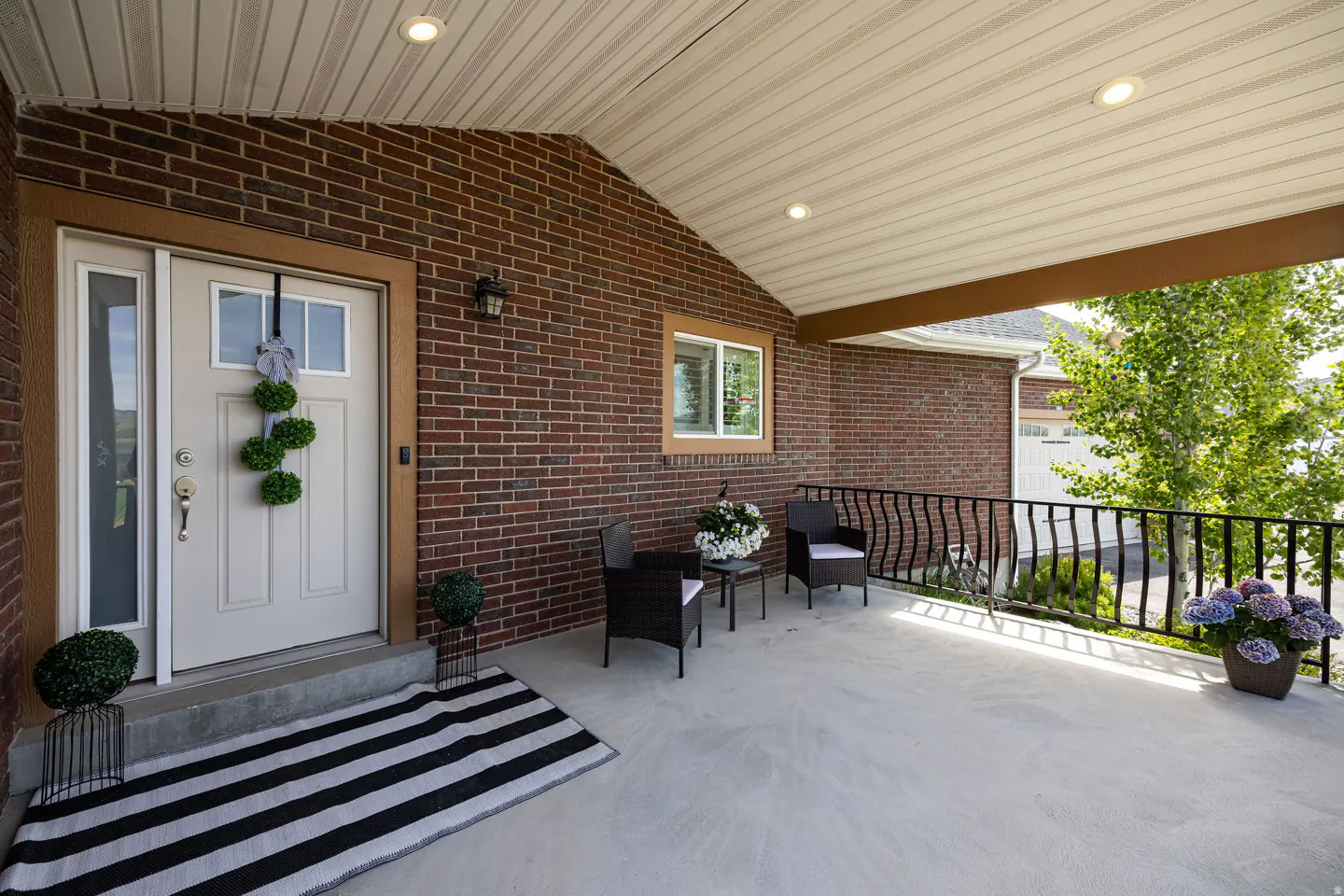 Covered porch of a brick house with a white door, striped rug, black chairs, and flowers.