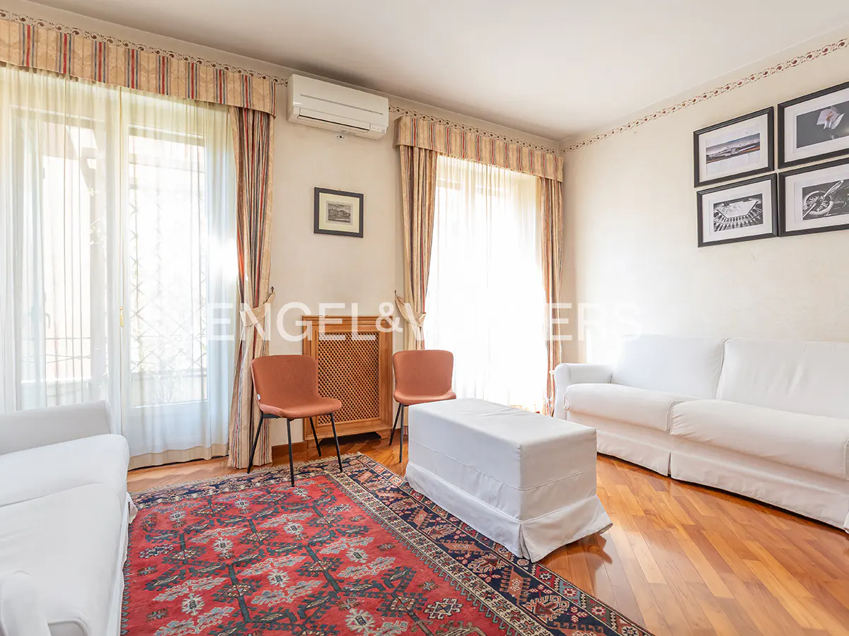 Bright living room with hardwood floors, white sofas, and a red patterned rug. Two orange chairs face a wooden radiator cover. Windows have striped valances.
