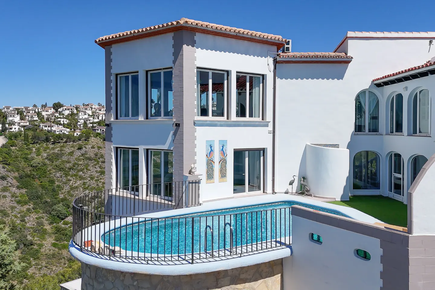 Exterior view of a white multi-story house with a blue pool and black metal railing. The house has many windows and a red tile roof.