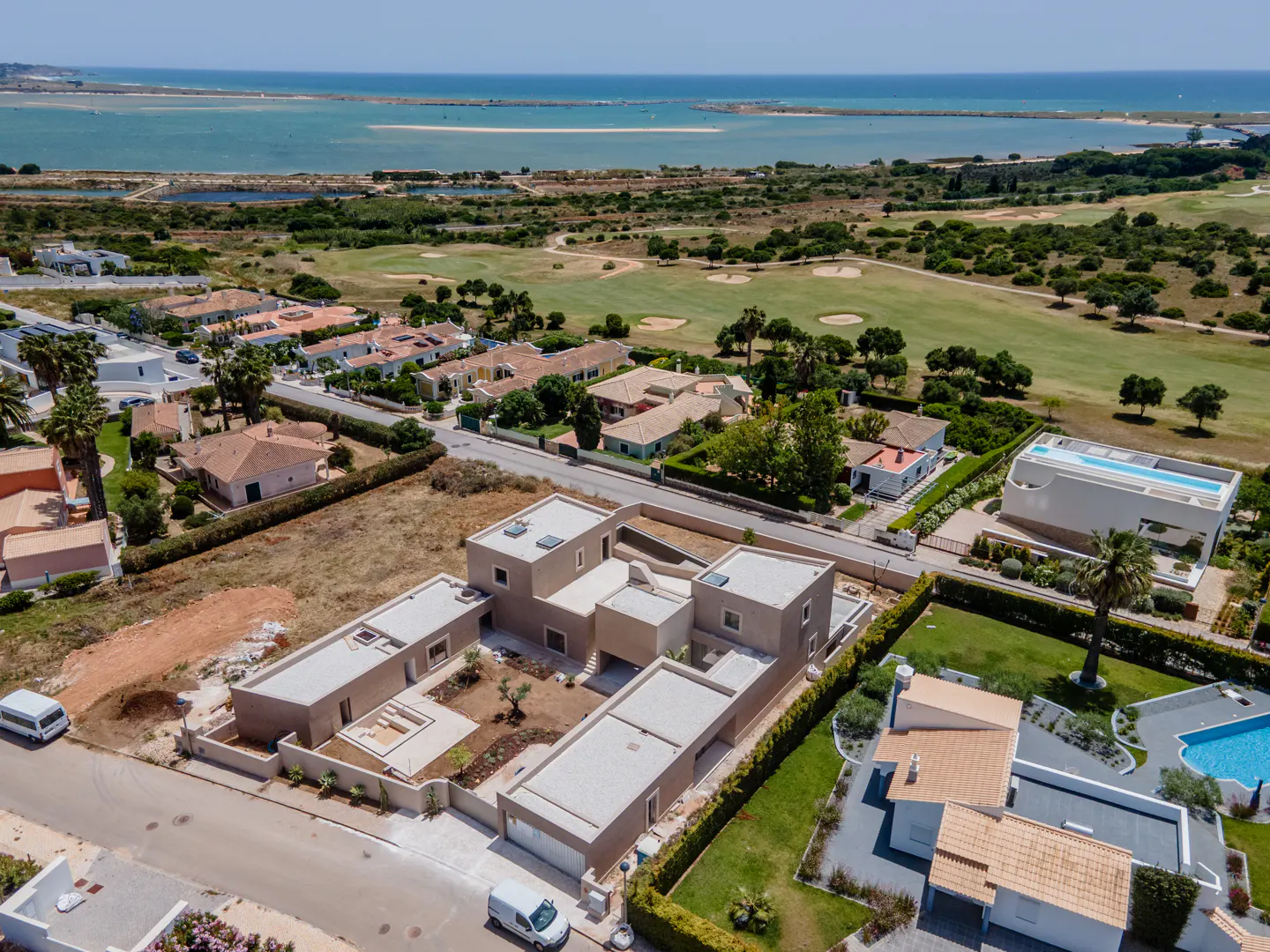Aerial view of a modern, beige house with a flat roof, surrounded by green lawns, other houses, and a golf course, with the ocean in the background.