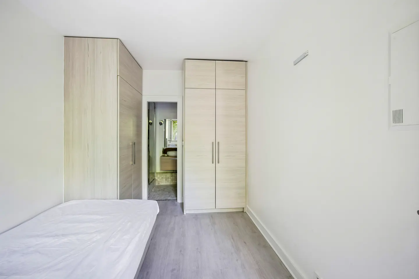 Bedroom with white walls, light wood wardrobes, and a bed with white sheets. A bathroom is visible through a doorway.