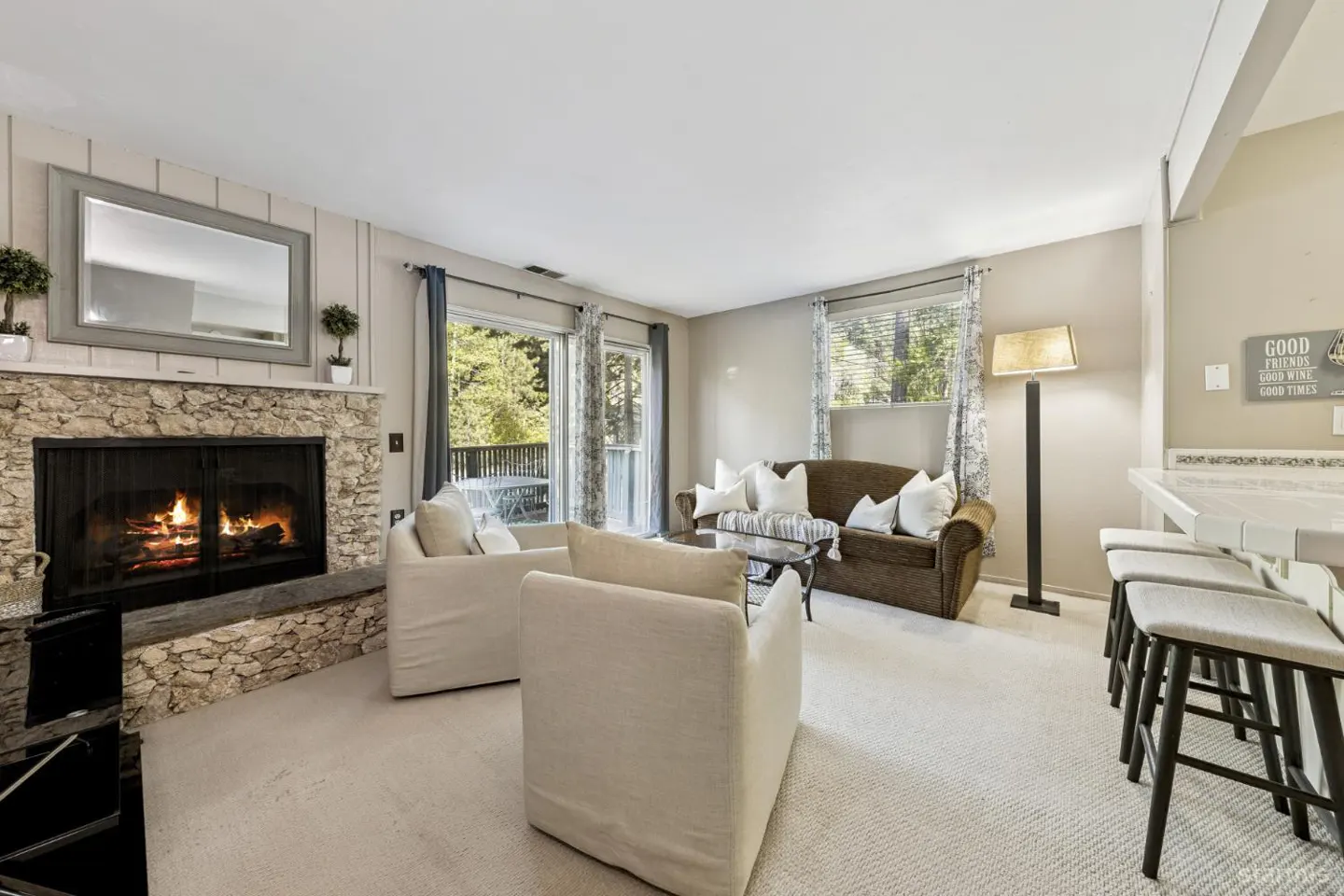 Living room with stone fireplace, white chairs, brown sofa, and bar stools. Fire is burning in the fireplace.