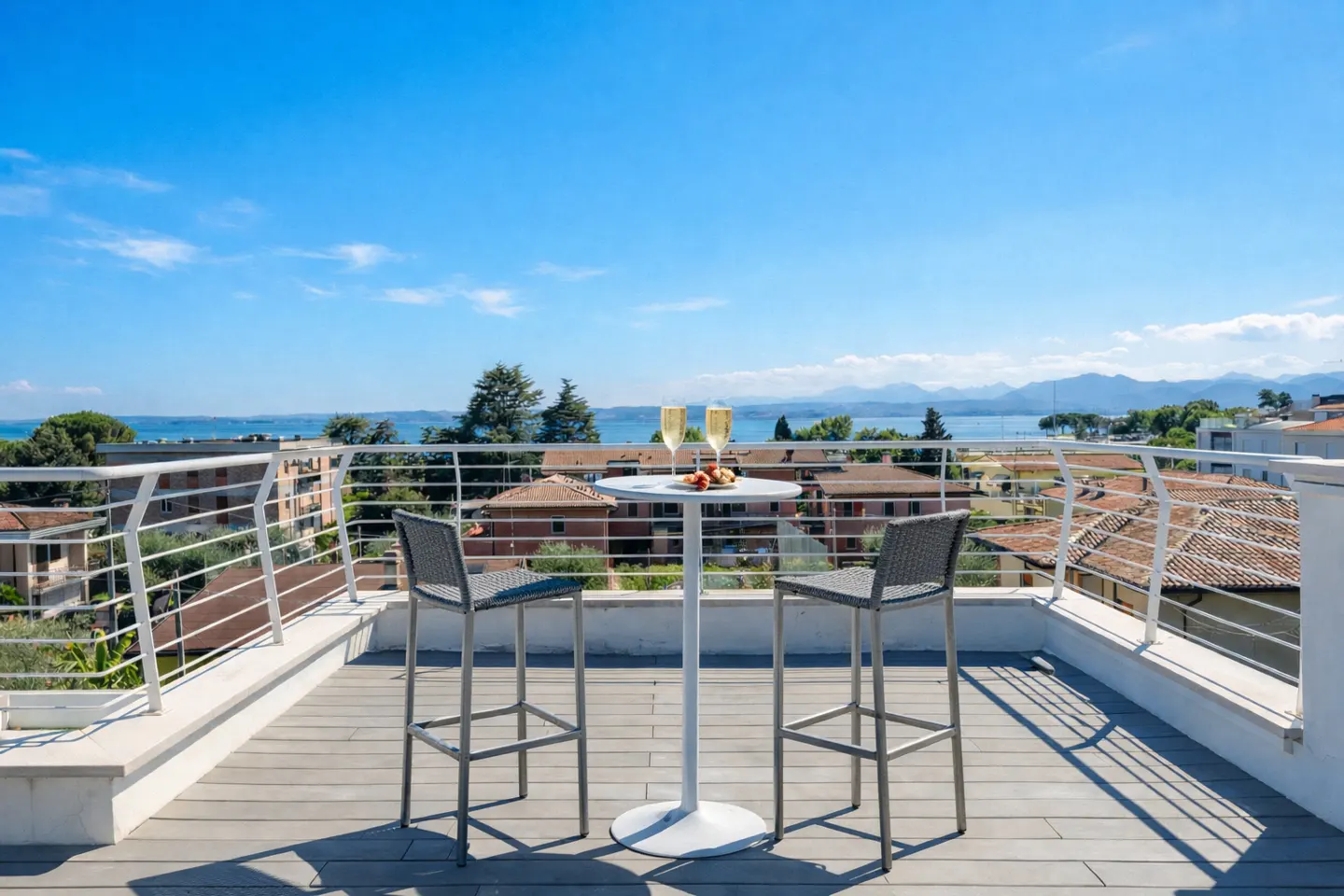 Rooftop patio with two gray bar stools, a white table, champagne flutes, and a scenic view of a lake and mountains.